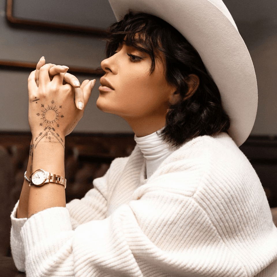 Woman wearing a white cowboy hat and sweater, with dark hair and henna tattoos on her hand.