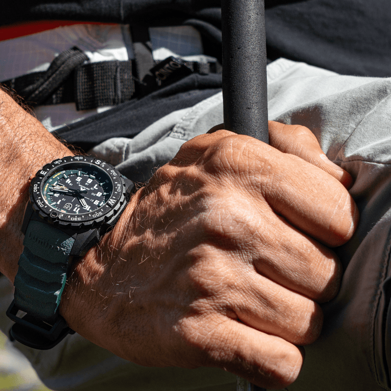 Wristwatch with a black dial and green strap worn on a hand gripping a pole.