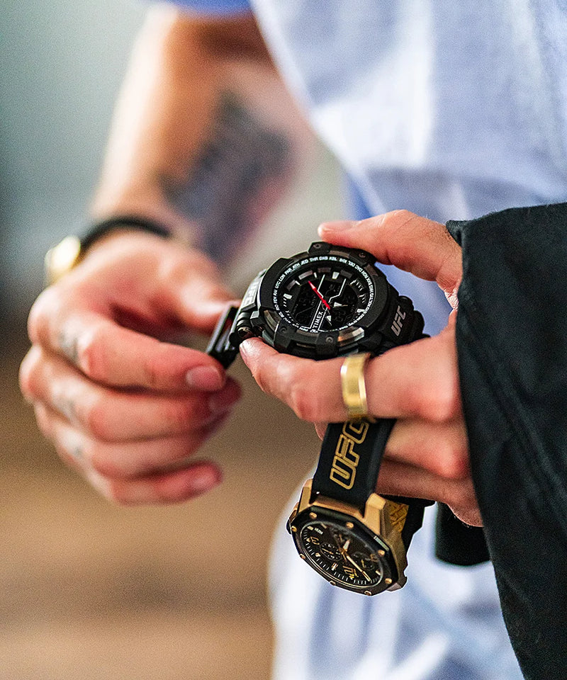 Wristwatch with a black face and gold-colored details being held in someone’s hands.