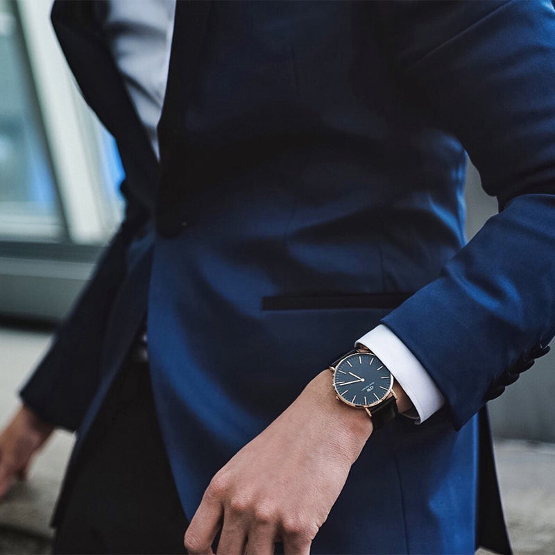 Elegant wristwatch with a dark face and slim leather strap on a person wearing a navy suit.